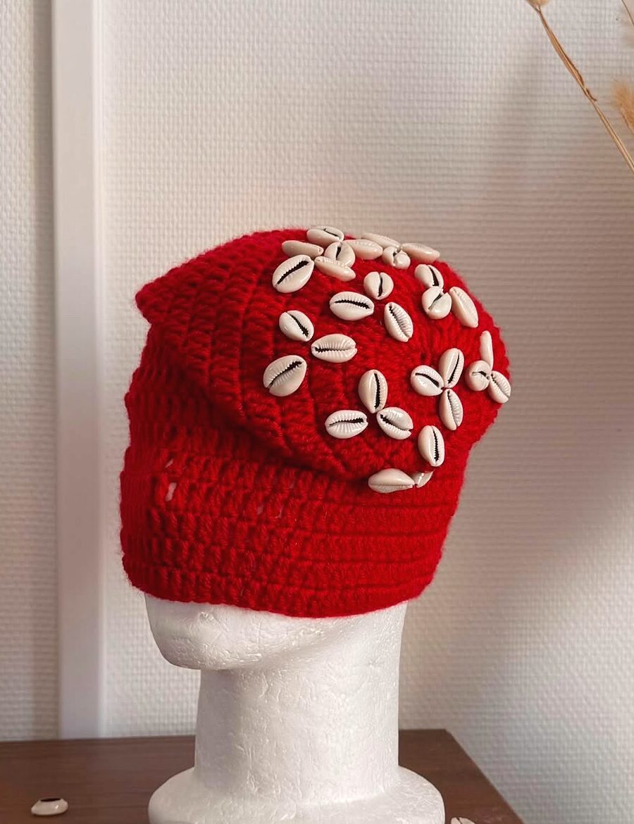 Bonnet Cauris — bonnet en crochet rouge parsemé de coquillages cauris, sur tête mannequin, fond clair