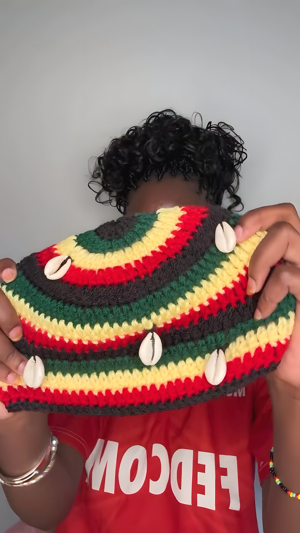 Bonnet fini à motif rasta et cauris présenté dans les mains de l'artisane