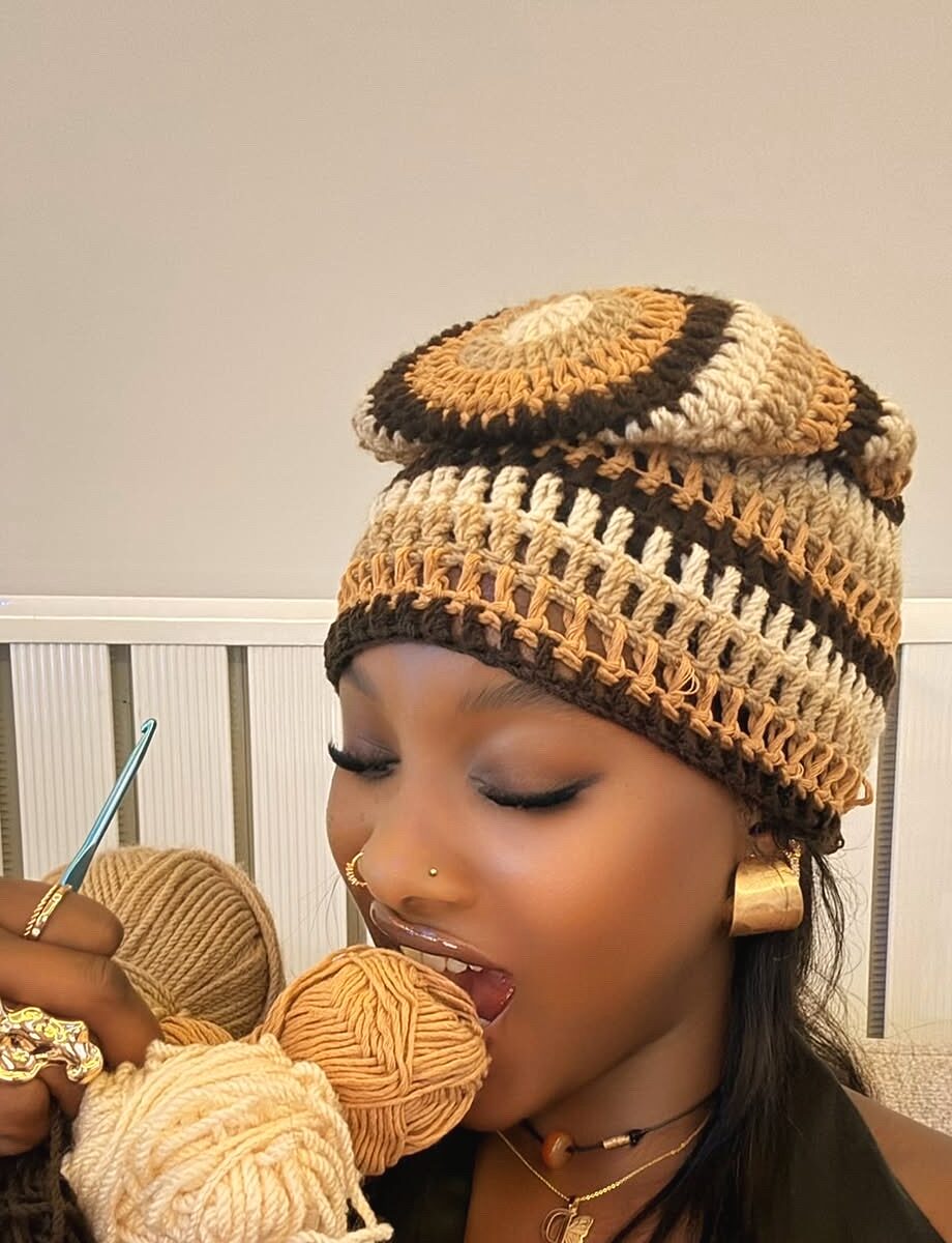 Muse coiffée d'un bonnet crocheté brun, tenant des pelotes de fil de coton et un crochet turquoise